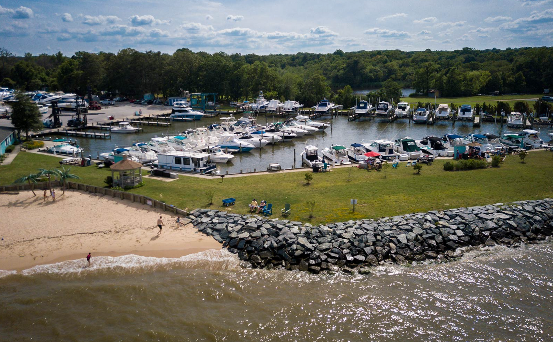 Boats docked at marina with sandy beach, green grass, trees, and cloudy sky.