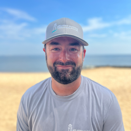 Man in gray hat and shirt smiles on a sandy beach, blurred ocean background.