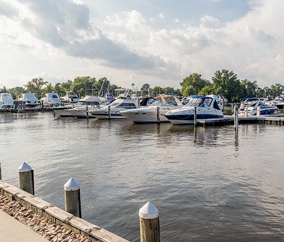 Boats docked at a marina on a cloudy day, wooden piers, calm water, and trees in the background.