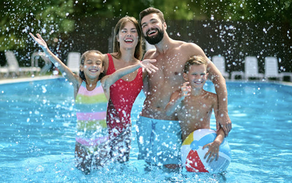 Family in a pool splashing water, smiling. Mom in red, kids, dad shirtless in blue shorts.
