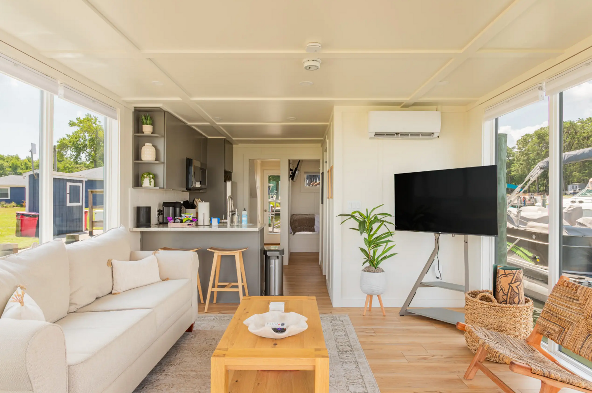 Living room in a white cottage with large windows. Beige couch, wooden coffee table, and kitchen area.