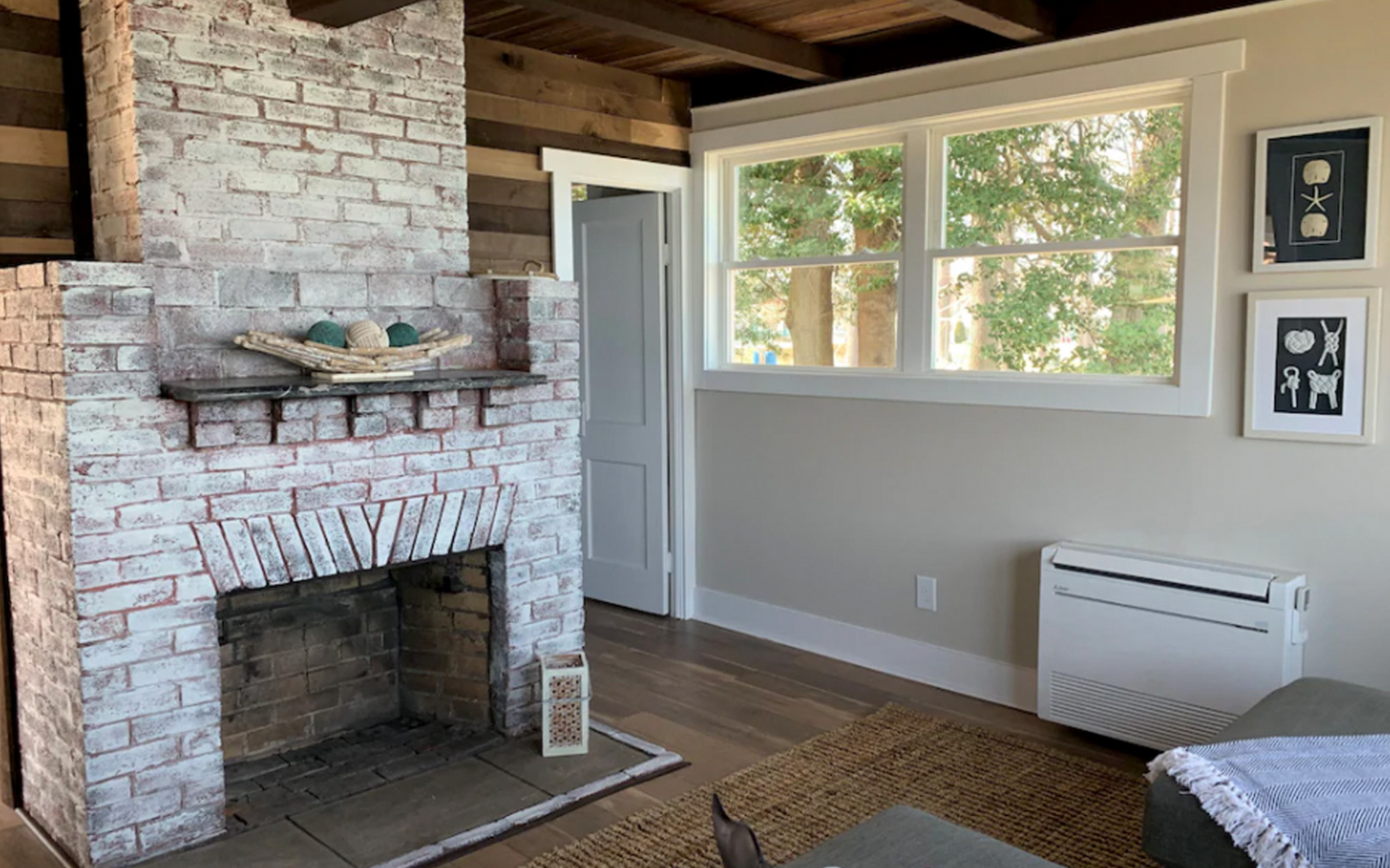 Cozy living room with whitewashed brick fireplace, wooden beams, large window, and framed art.
