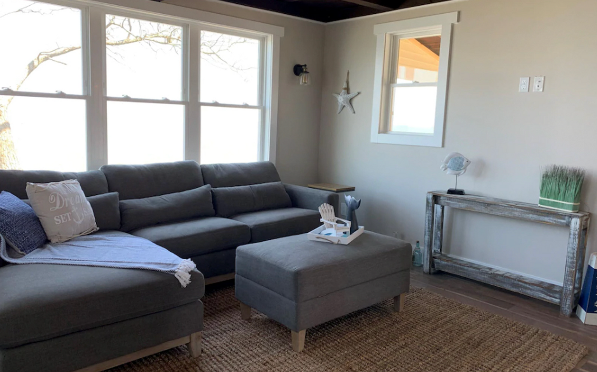 Living room with gray sectional sofa, ottoman, and rustic wood console table.
