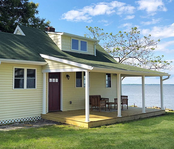 Yellow house with green roof and a porch overlooking a body of water under a blue sky.
