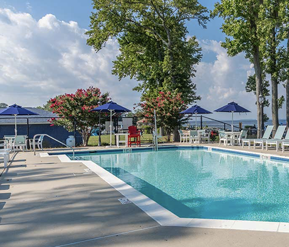 Pool with blue water, lounge chairs, umbrellas, and trees under a cloudy sky.