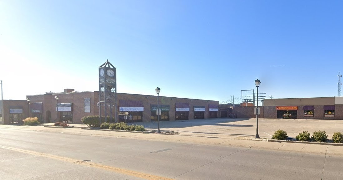 A strip mall with a clock tower under a clear blue sky. Empty parking area in front.