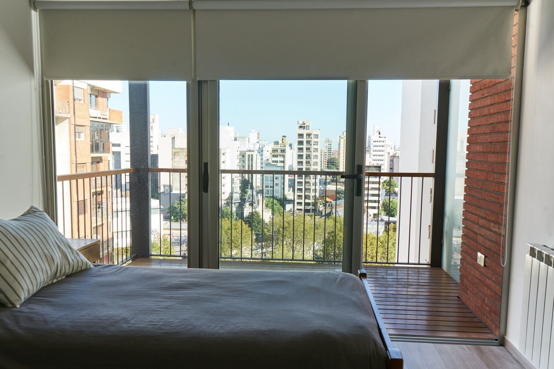 Dormitorio con cama frente a puertas abiertas con vista a la ciudad, cortinas enrollables color beige claro, pared de ladrillo.