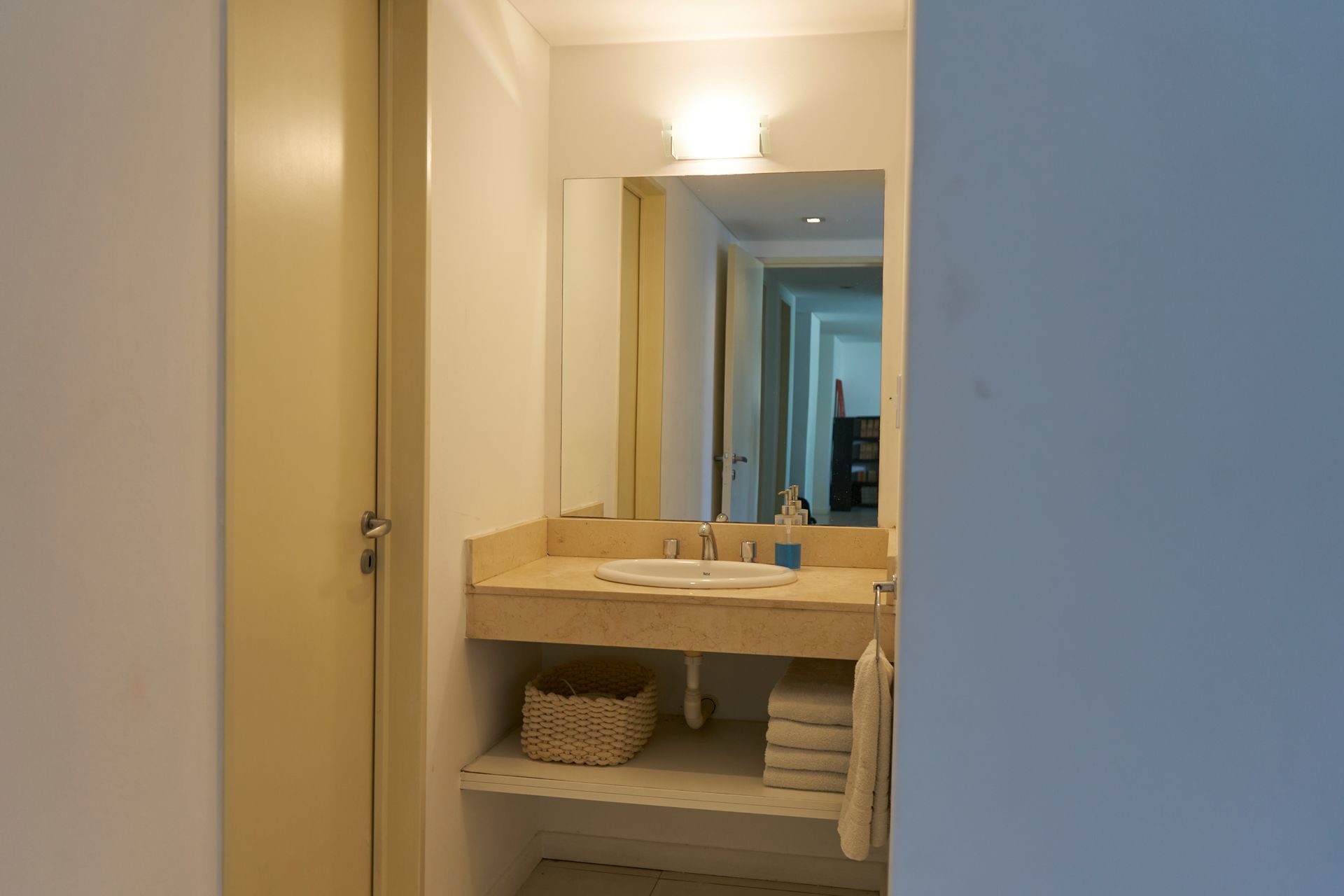 Baño con encimera de color beige, lavabo, espejo y cesta tejida.