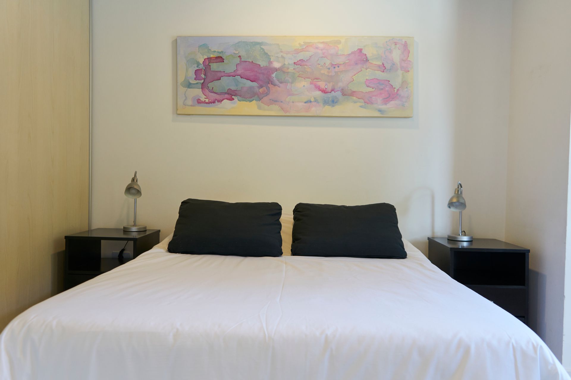 Dormitorio con una cama, dos almohadas negras, mesitas de noche, lámparas y arte abstracto.