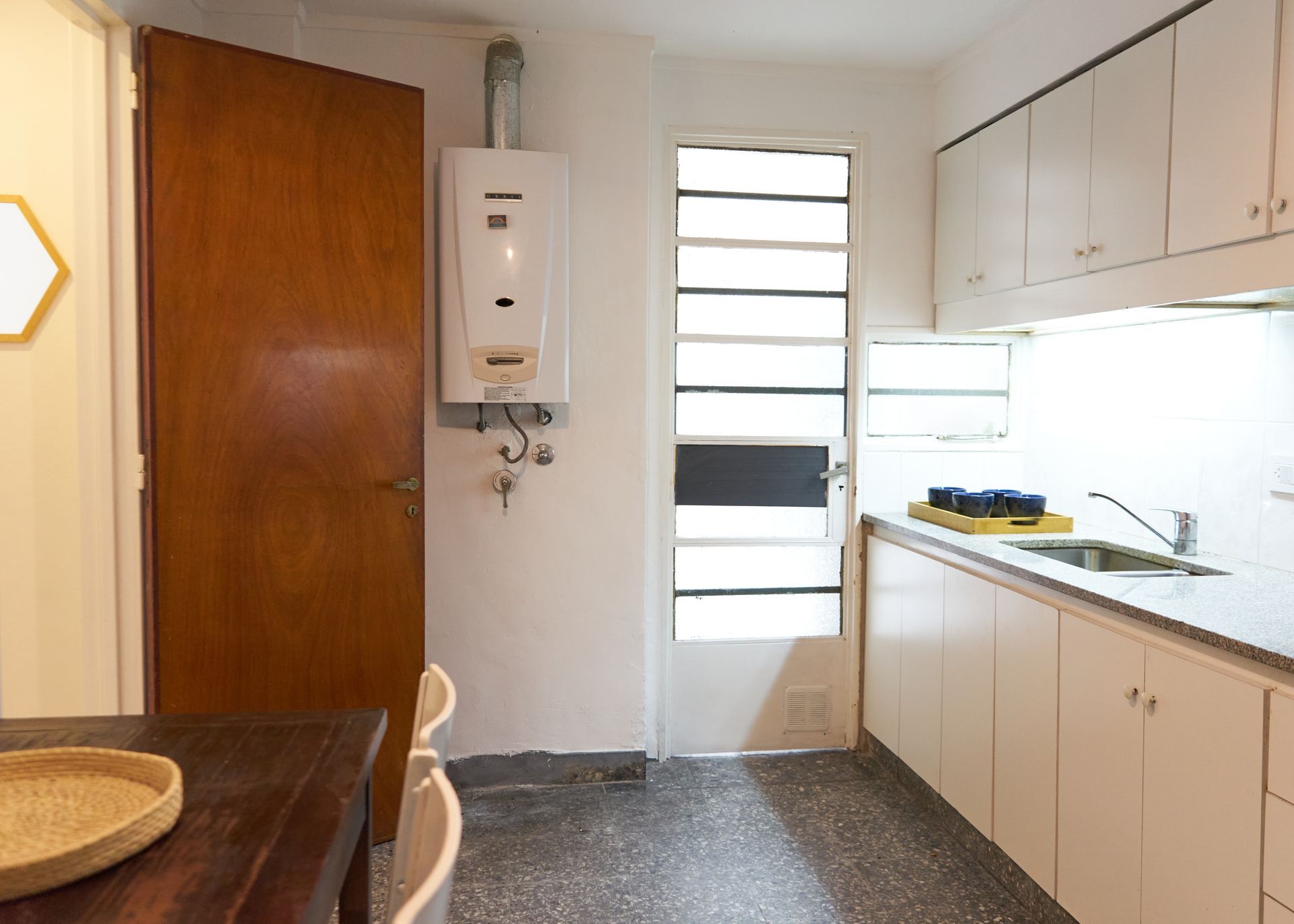 Cocina pequeña con calentador de agua, gabinetes blancos, puerta marrón cerrada y ventana.