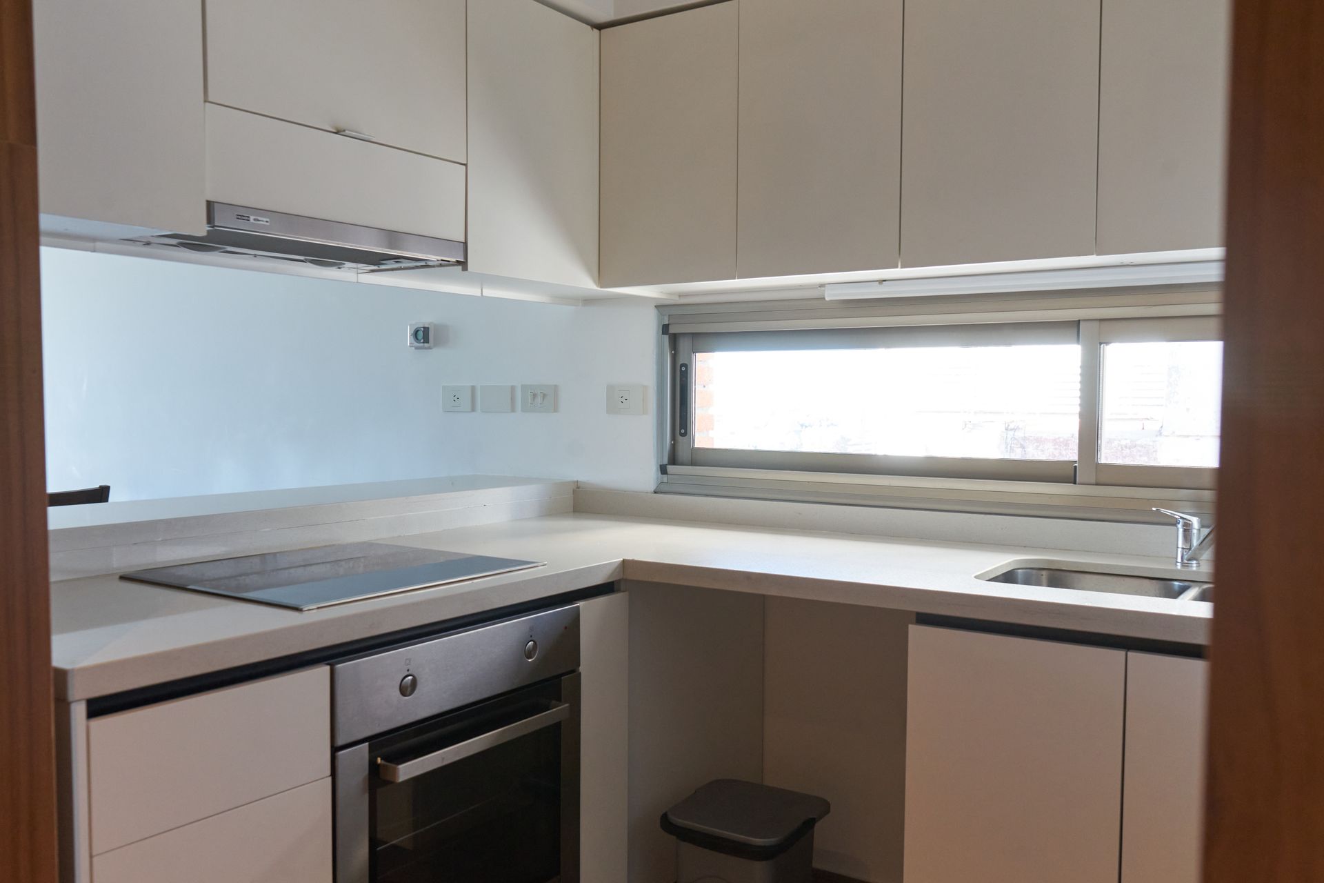 Cocina moderna y blanca con horno empotrado, placa de cocina, fregadero y armarios altos. Se ven una pequeña ventana y un cubo de basura.