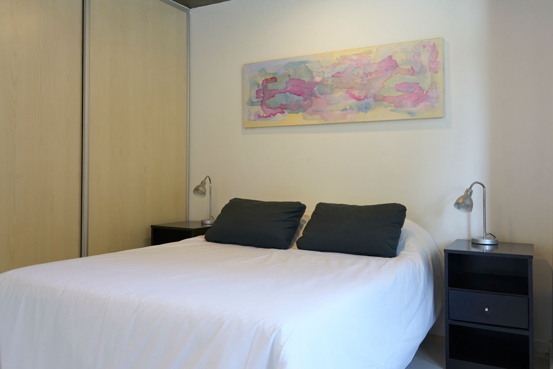 Dormitorio con ropa de cama blanca, almohadas y mesitas de noche negras. Una obra de arte en tonos claros cuelga sobre la cama.