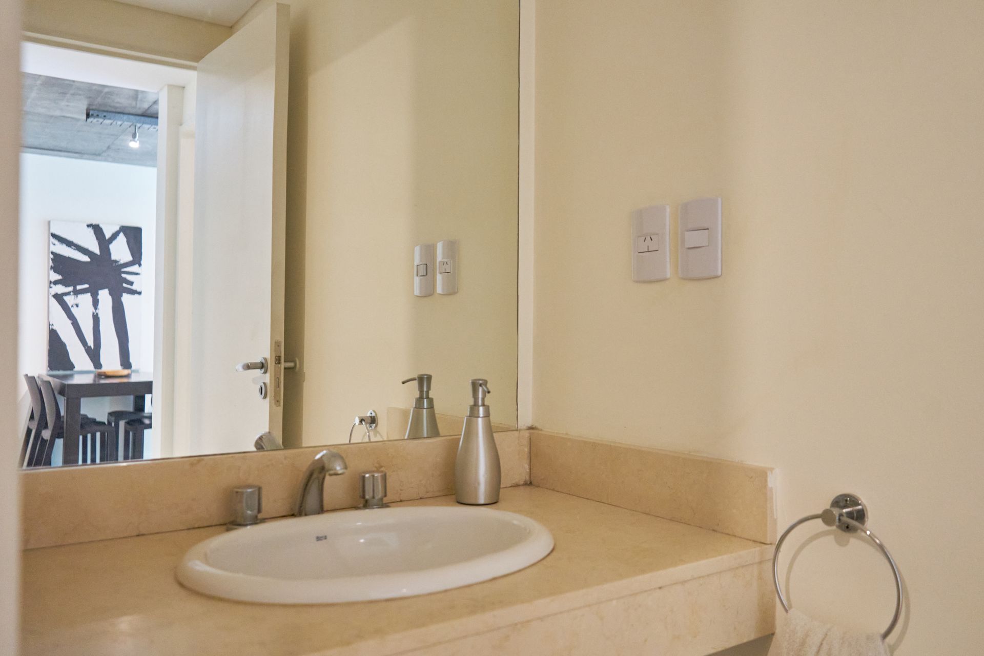 Baño con encimera color crema, lavabo blanco y espejo grande. Pared color crema claro. Hay una toalla colgada en la pared.