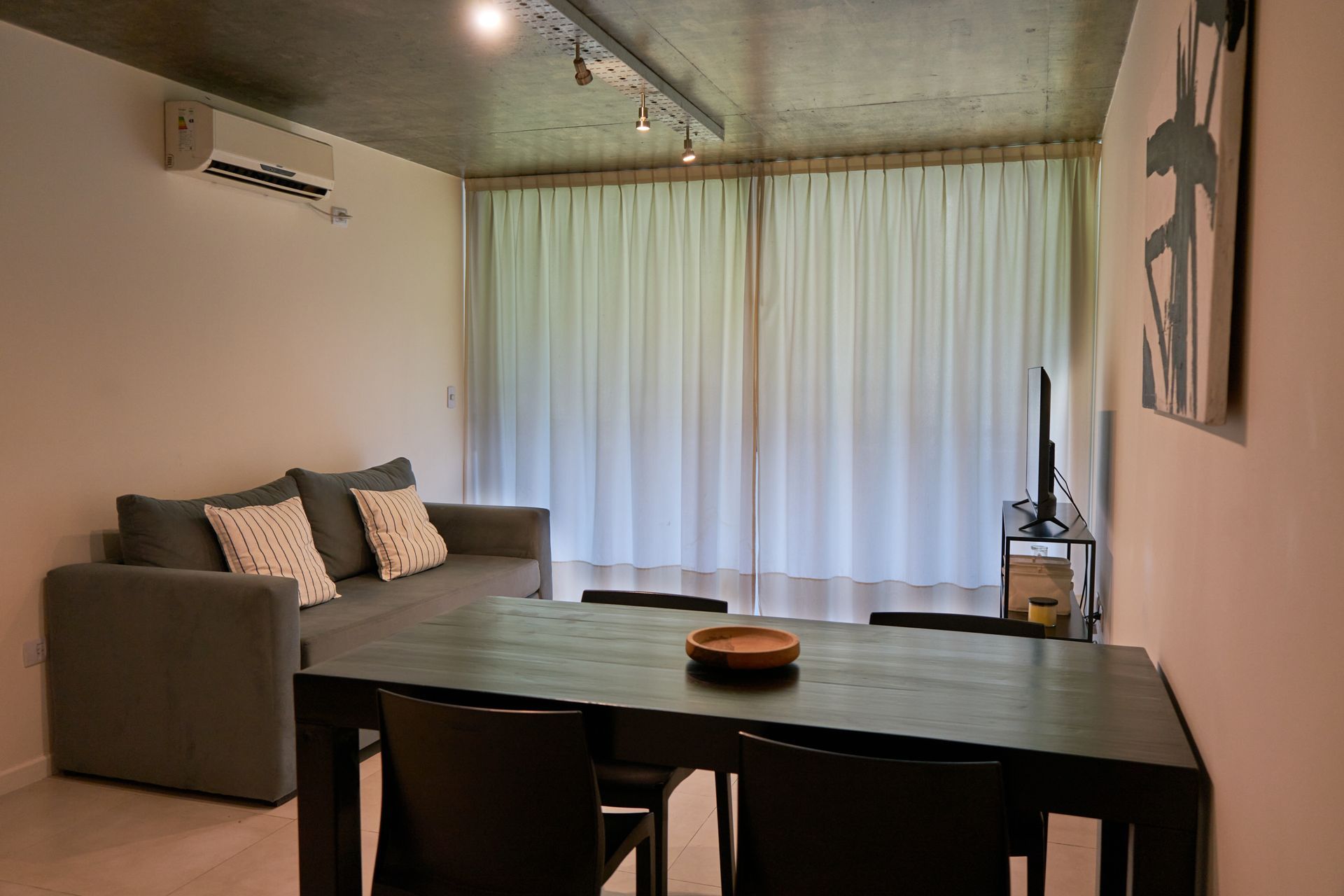 Sala de estar con sofá gris, mesa de comedor de madera oscura, cortinas blancas y TV.
