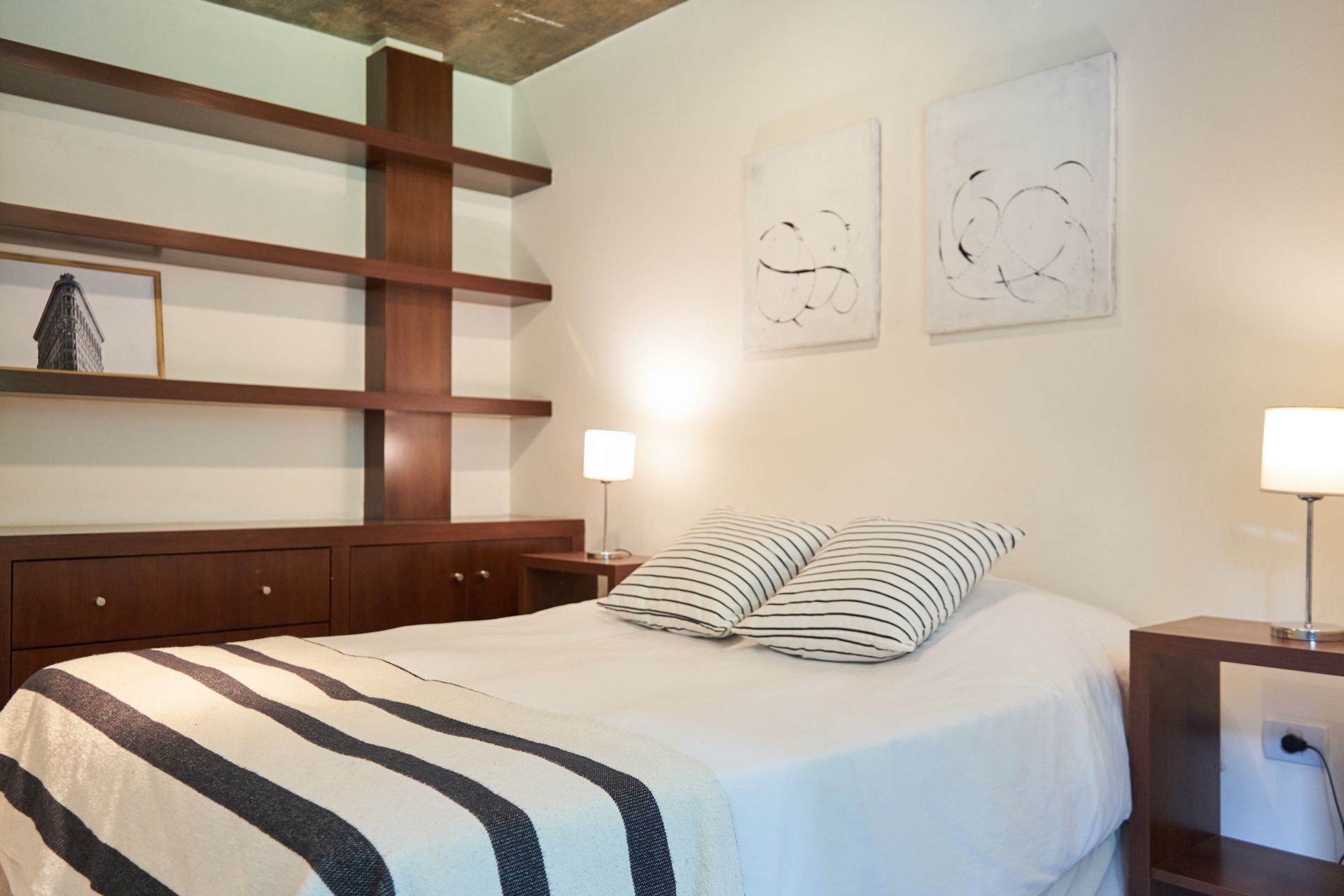 Dormitorio con cama, estanterías empotradas y obras de arte. Paredes blancas, muebles de madera y dos lámparas.