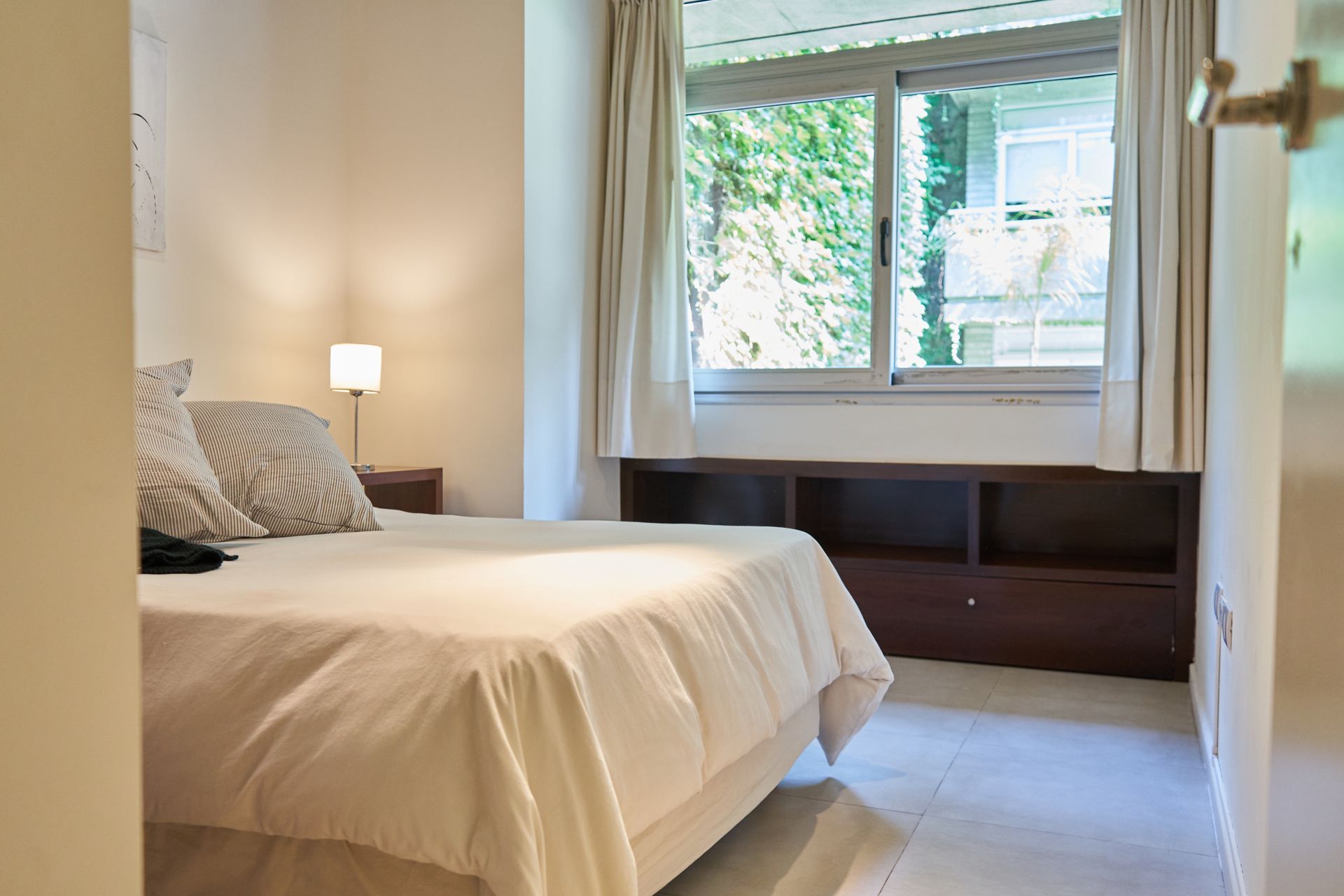 Dormitorio con cama, almohadas, mesita de noche, ventana con vista a los árboles y almacenamiento incorporado.