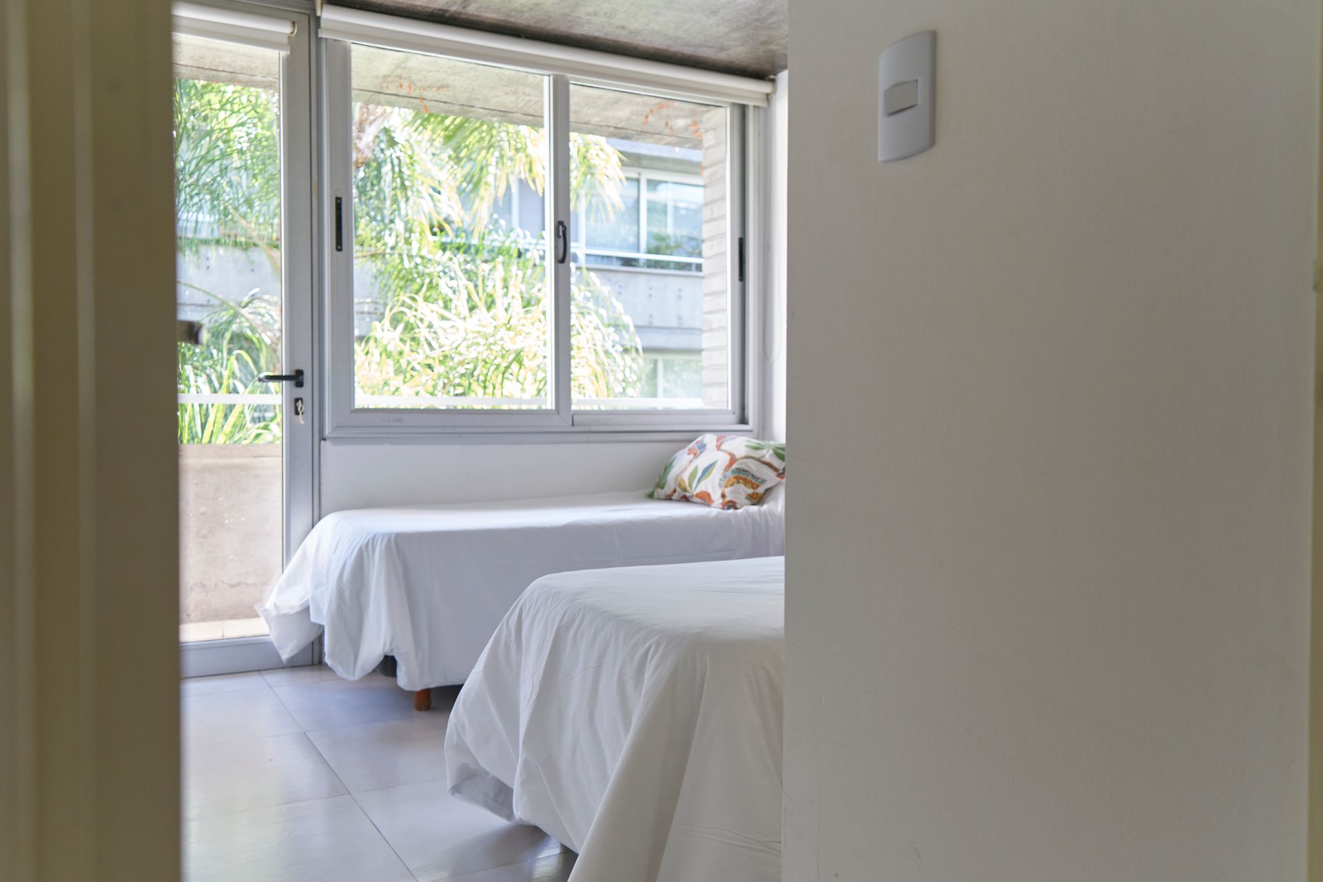 Dos camas con sábanas blancas cerca de una ventana, visible desde una puerta.