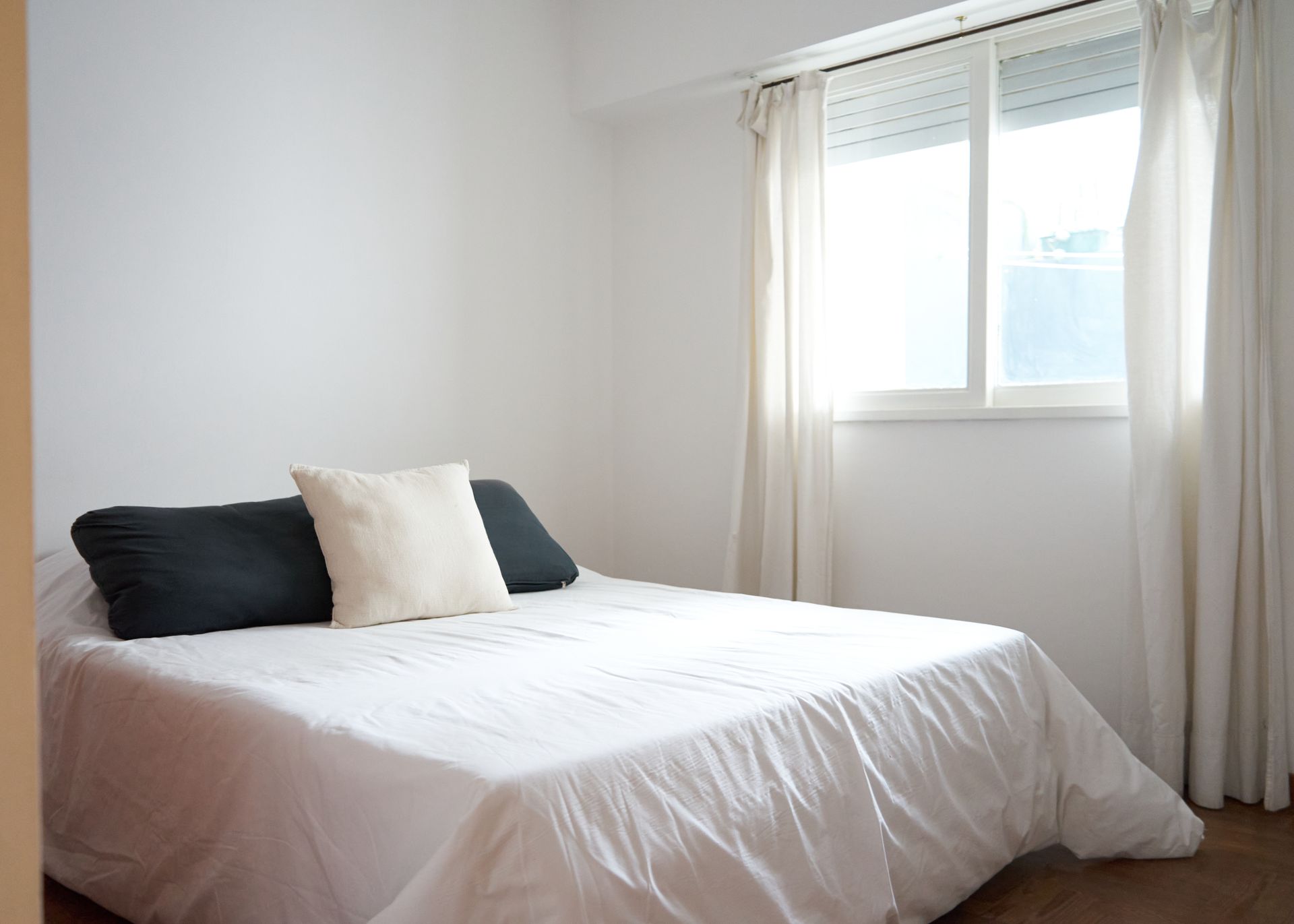 Dormitorio blanco con cama, almohadas, ventana y cortinas.