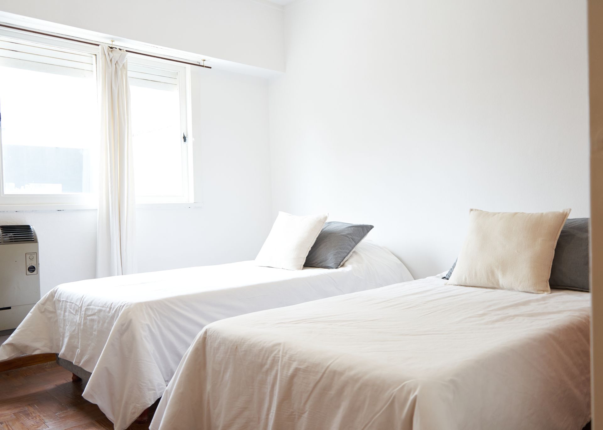 Un dormitorio blanco luminoso con dos camas individuales, sábanas blancas y una ventana con cortinas.