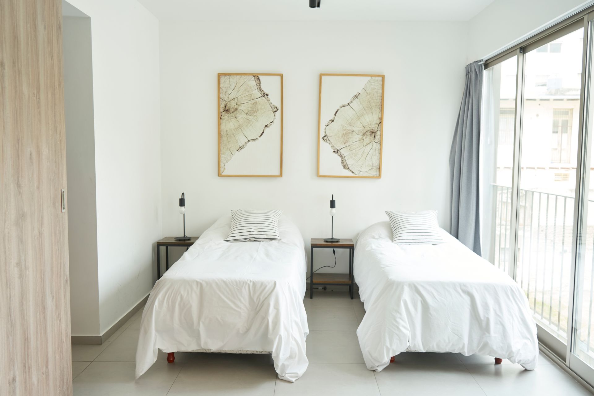 Dormitorio con dos camas individuales, ropa de cama blanca, láminas artísticas y acceso al balcón.