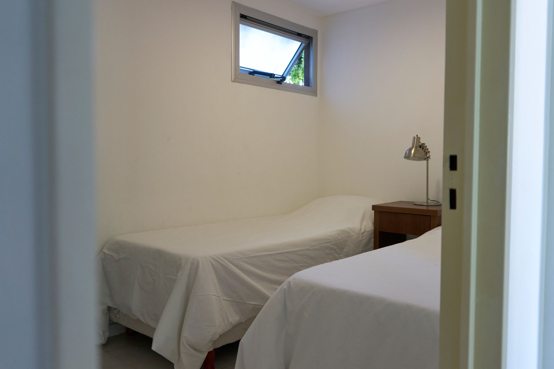 Dos camas individuales en una habitación pequeña con una pequeña ventana y una lámpara en una mesita de noche.