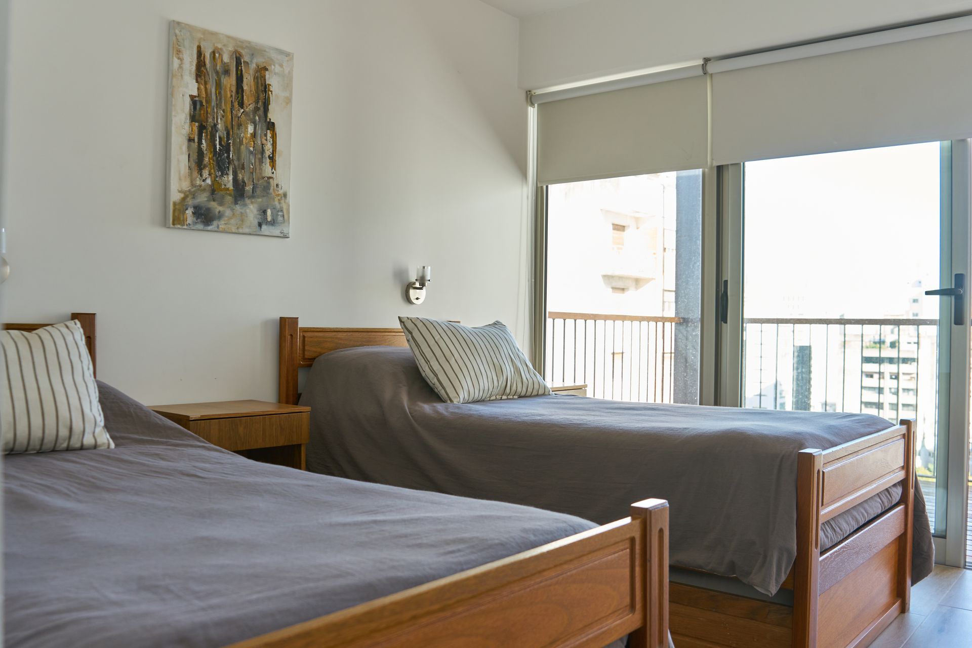 Dos camas individuales en una habitación luminosa con vista al balcón y un cuadro abstracto en la pared.