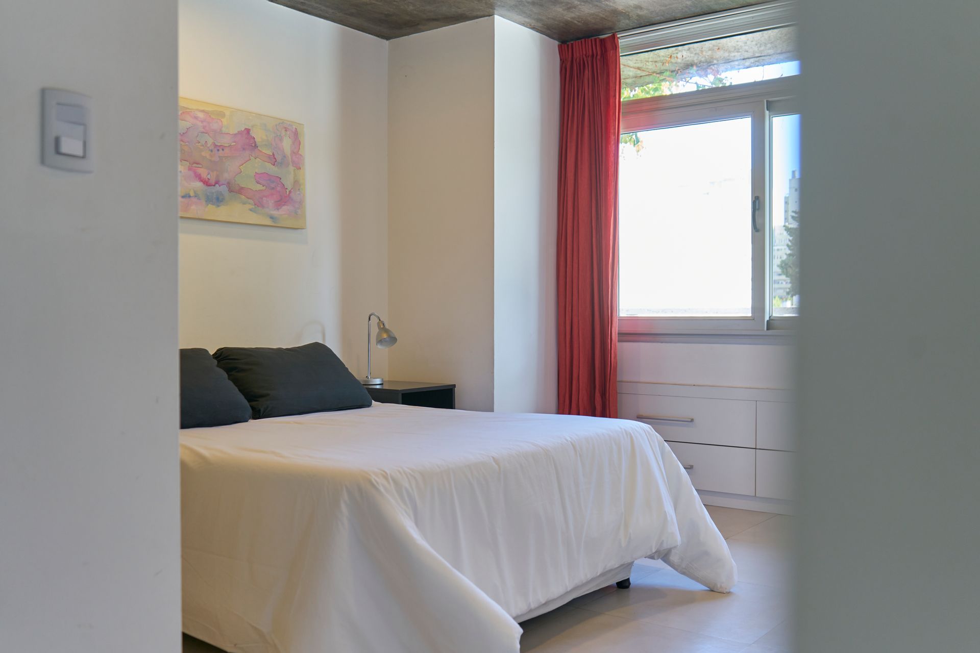 Dormitorio con cama blanca, almohadas gris oscuro, cortina roja y obras de arte en la pared.