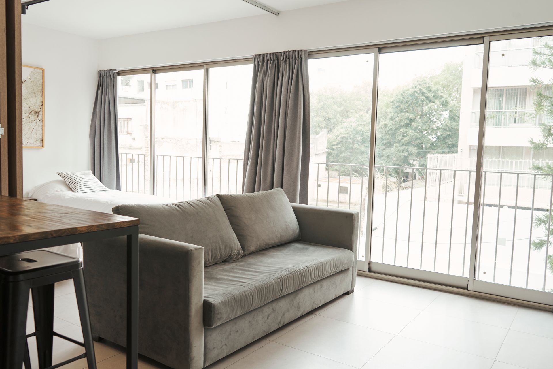 Pequeño apartamento con un sofá gris, grandes ventanales y una cama al fondo.
