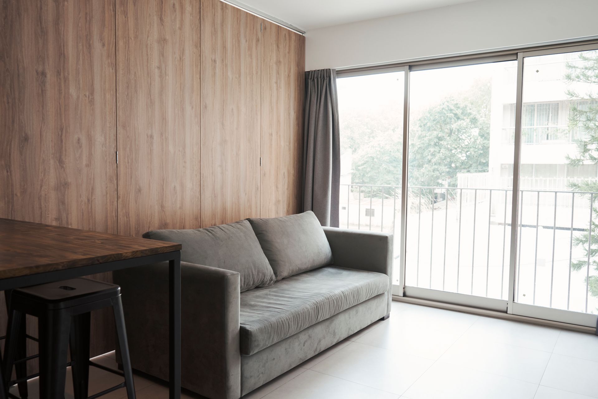 Pequeño espacio de estar con pared de madera, sofá gris y ventana grande.