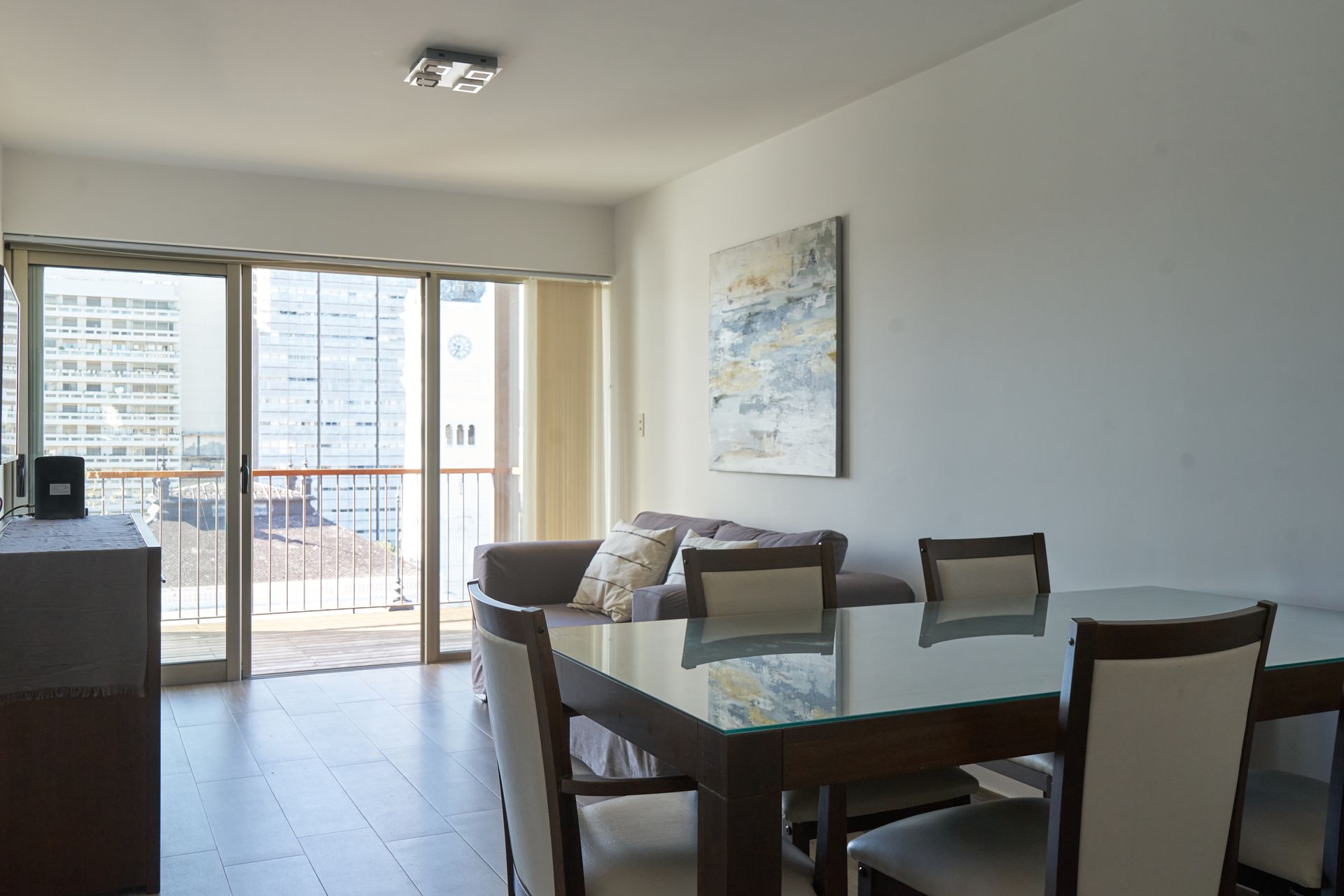 Sala de estar con mesa de comedor de vidrio, sofá, balcón y vista al paisaje urbano.