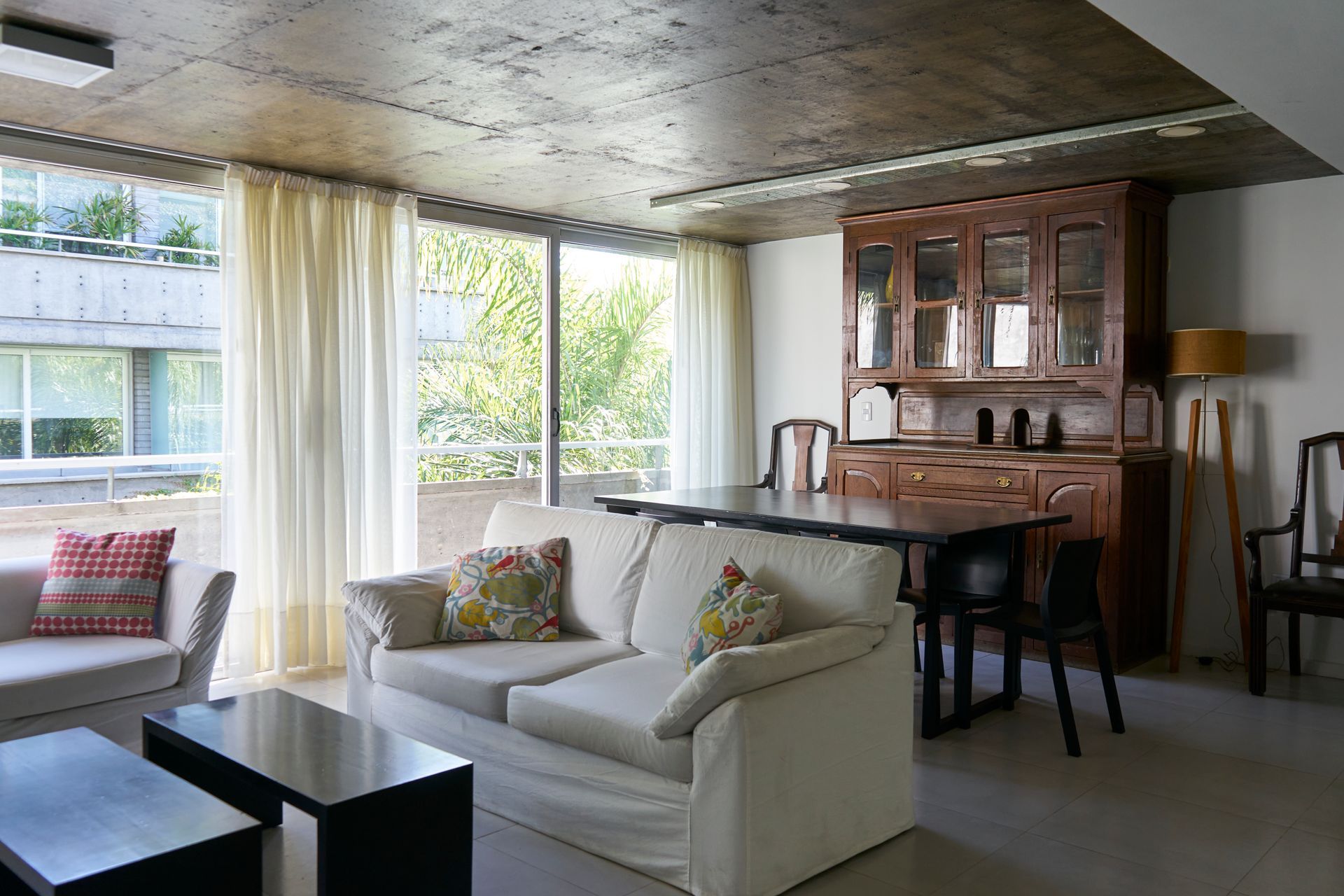 Sala de estar con sofá color crema, mesa oscura, mueble de madera y grandes ventanales.