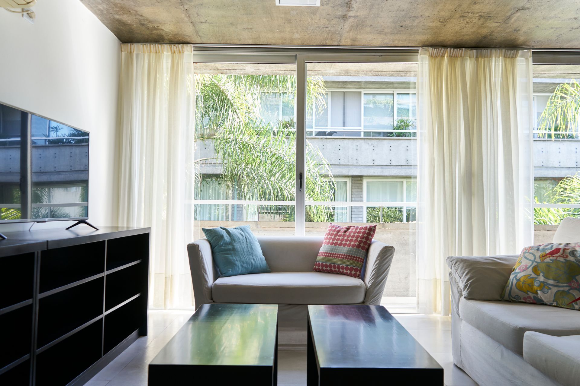 Sala de estar con sofá blanco, almohadas azules y estampadas, mesas de café negras y ventana alta con cortinas transparentes, vista exterior del edificio.