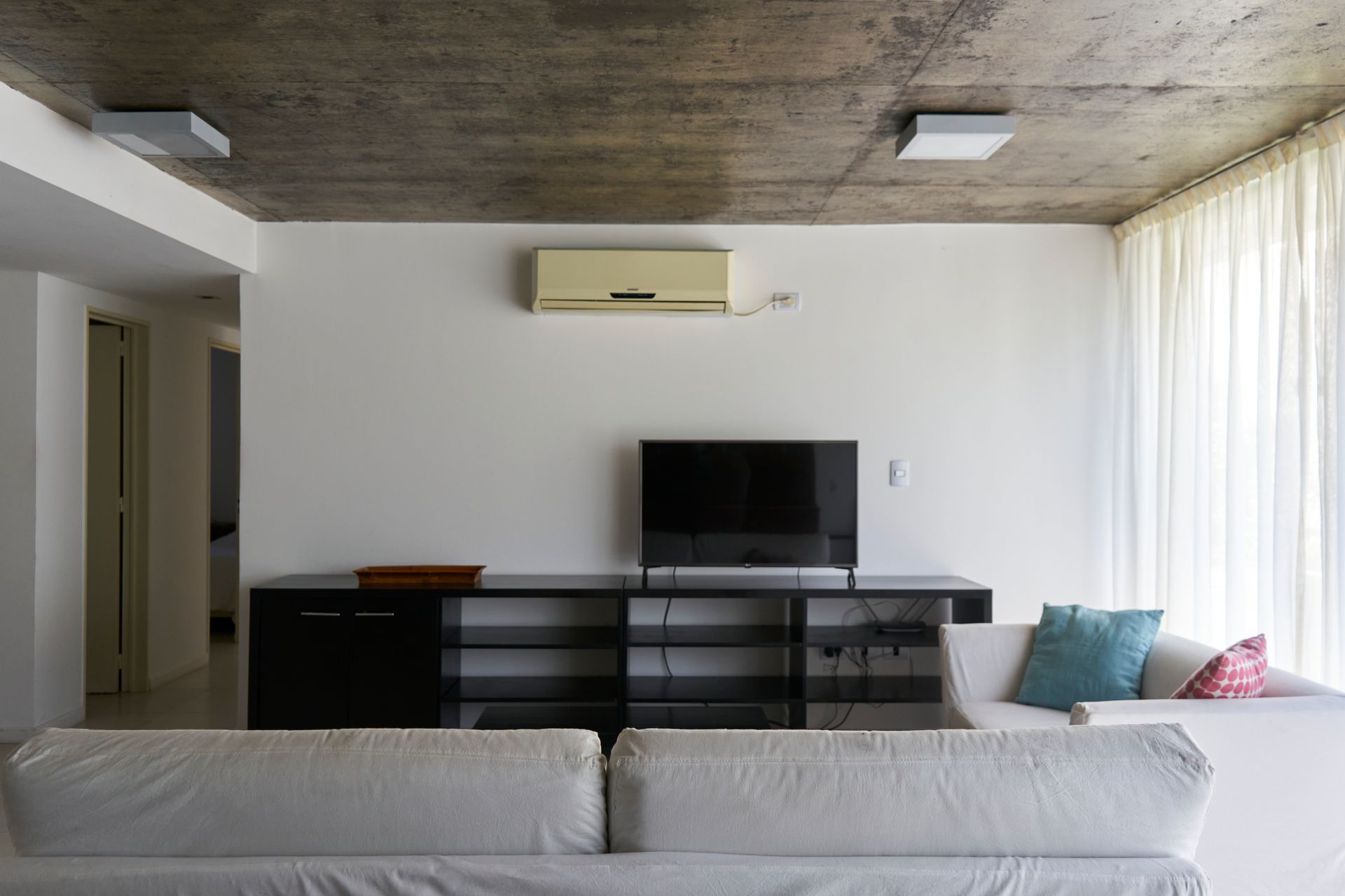 Sala de estar con sofá blanco, TV, centro de entretenimiento y techo de concreto.
