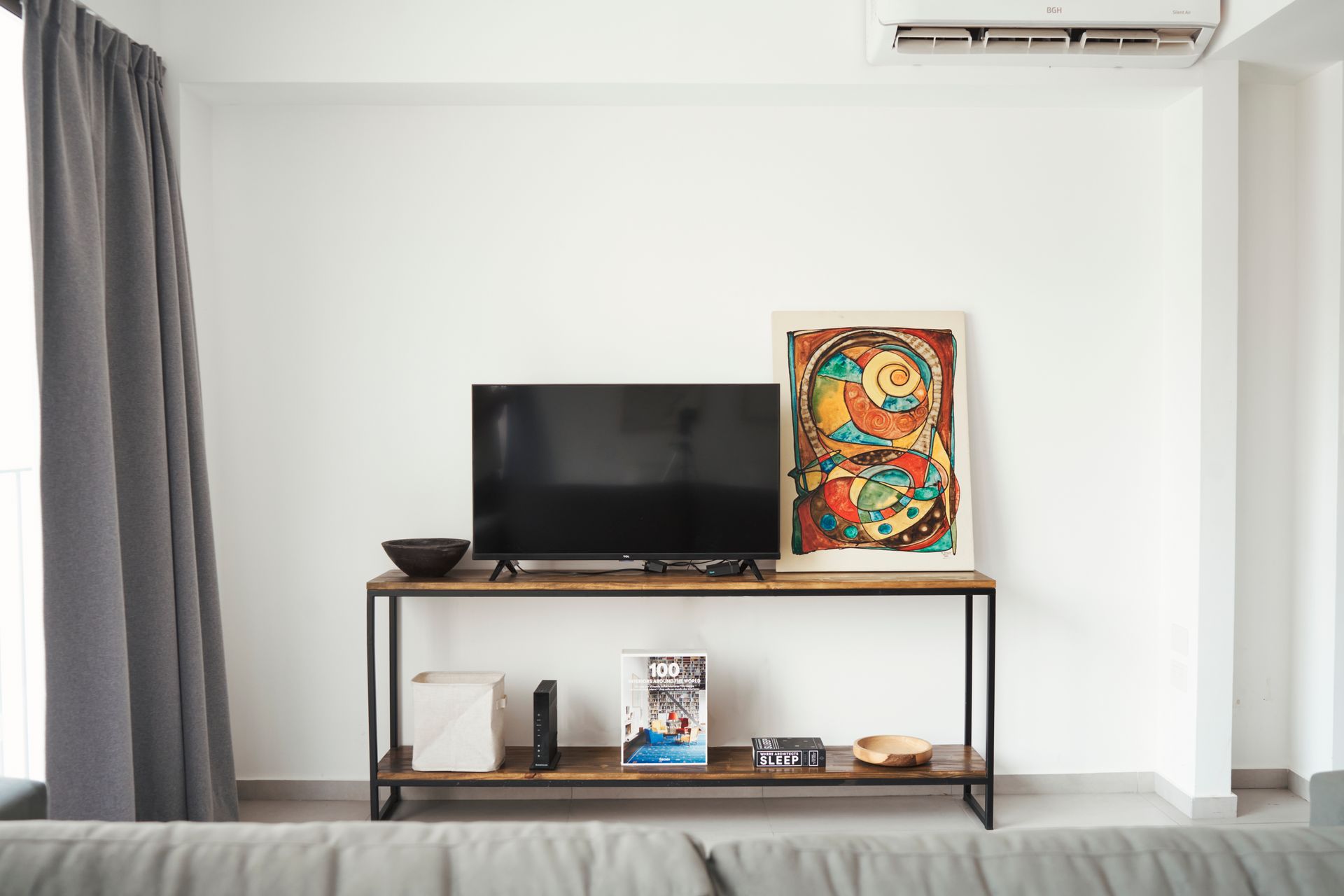 Sala de estar con TV en consola de madera, obras de arte, cortinas grises y aire acondicionado.