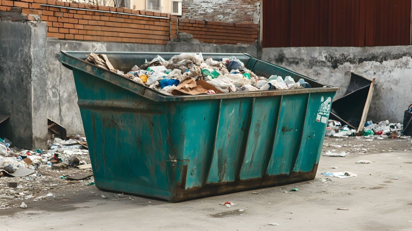 Overflowing green dumpster in a trash-strewn alley beside a brick wall