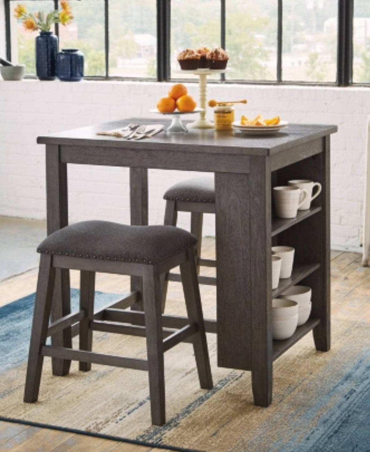 Gray kitchen island with stools, shelves holding cups, food on top, near window.