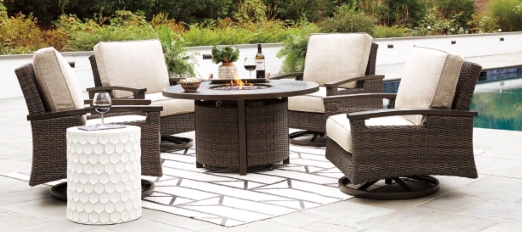 Outdoor wicker seating set with fire pit on a patio. Cream cushions, dark brown wicker, white side table, pool background.