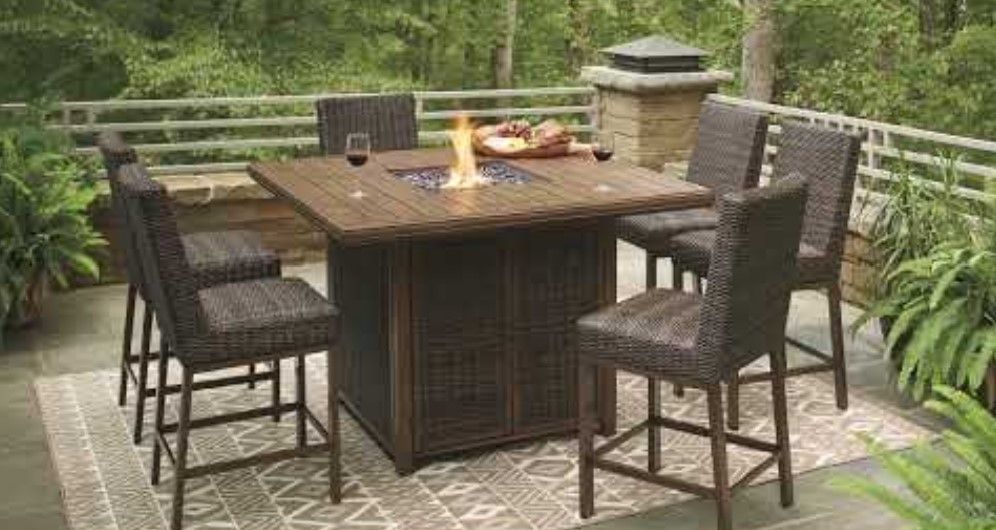 Outdoor dining set: brown wicker chairs around a square fire pit table on a patio.