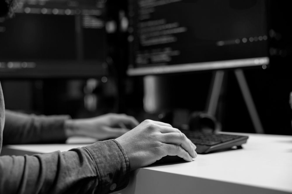 A person’s hands resting on a computer mouse and keyboard in front of monitors displaying code in a dark office.