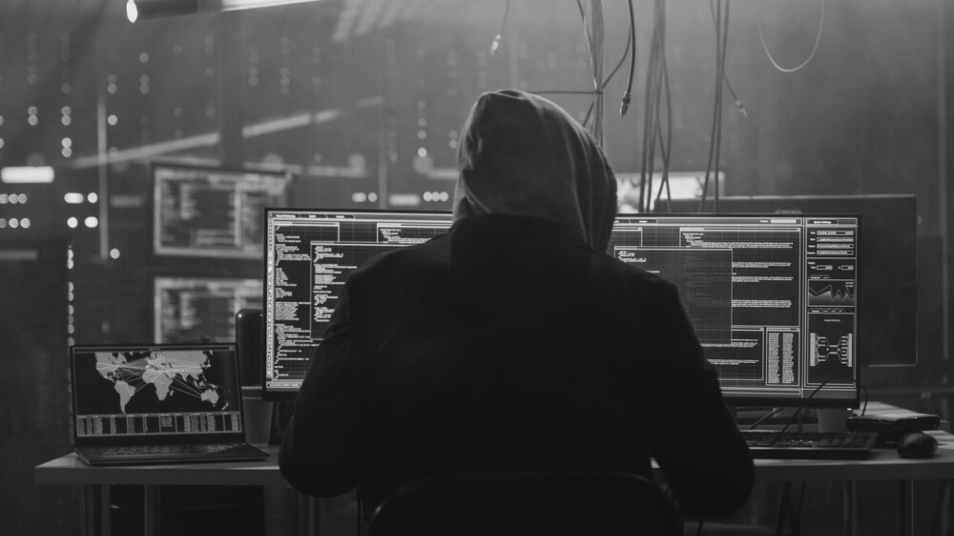 A man in a hood is sitting in front of a computer in a dark room.