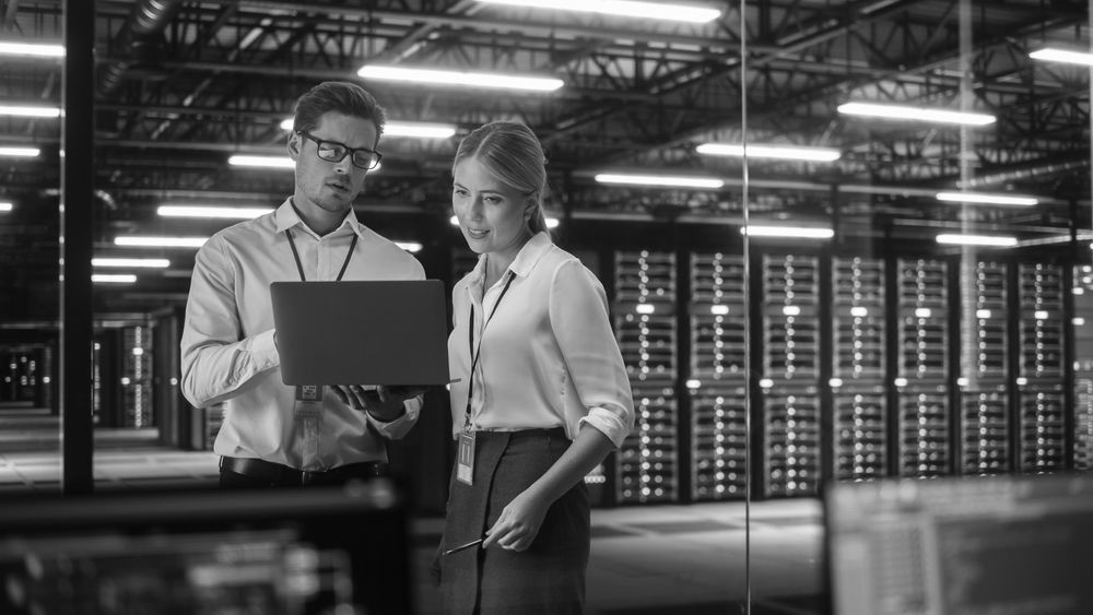 Two people in a server room looking at a laptop.