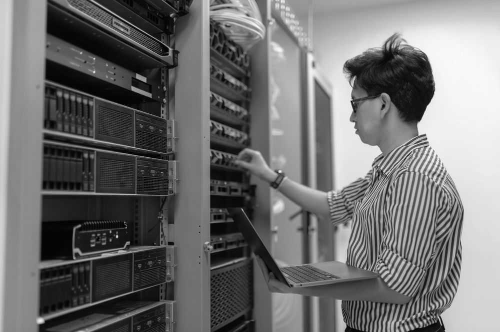 A man is working on a laptop in a server room.