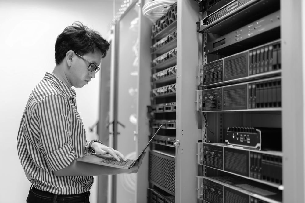 A man is working on a laptop in a server room.