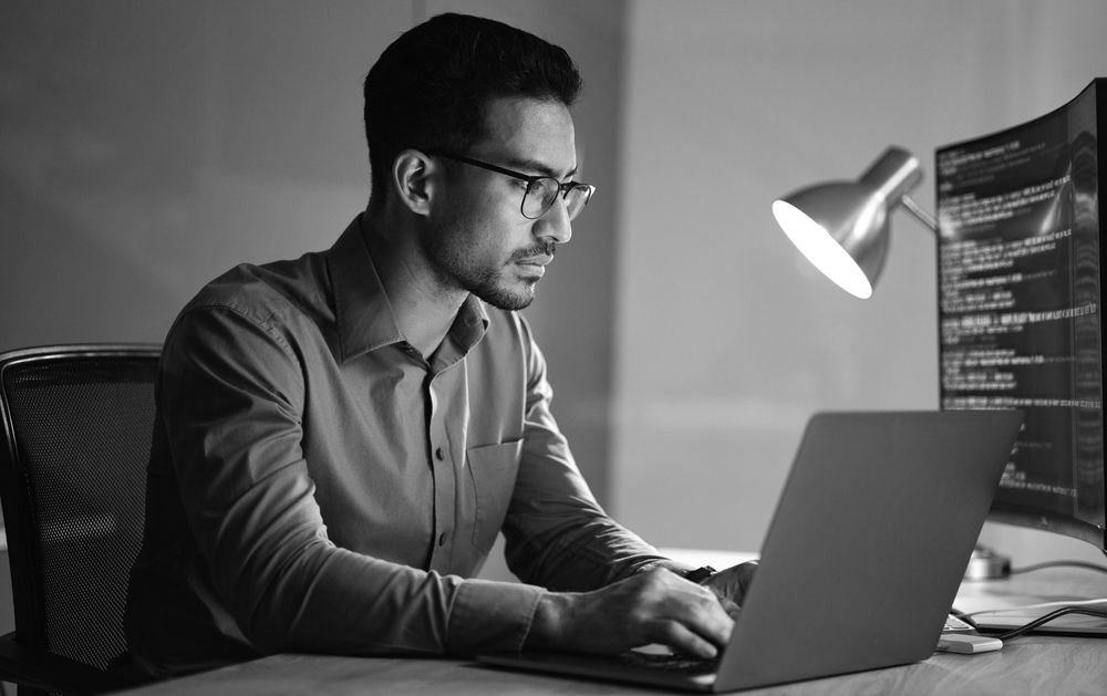 A focused individual wearing glasses works on a laptop in a dimly lit office with code displayed on a monitor.