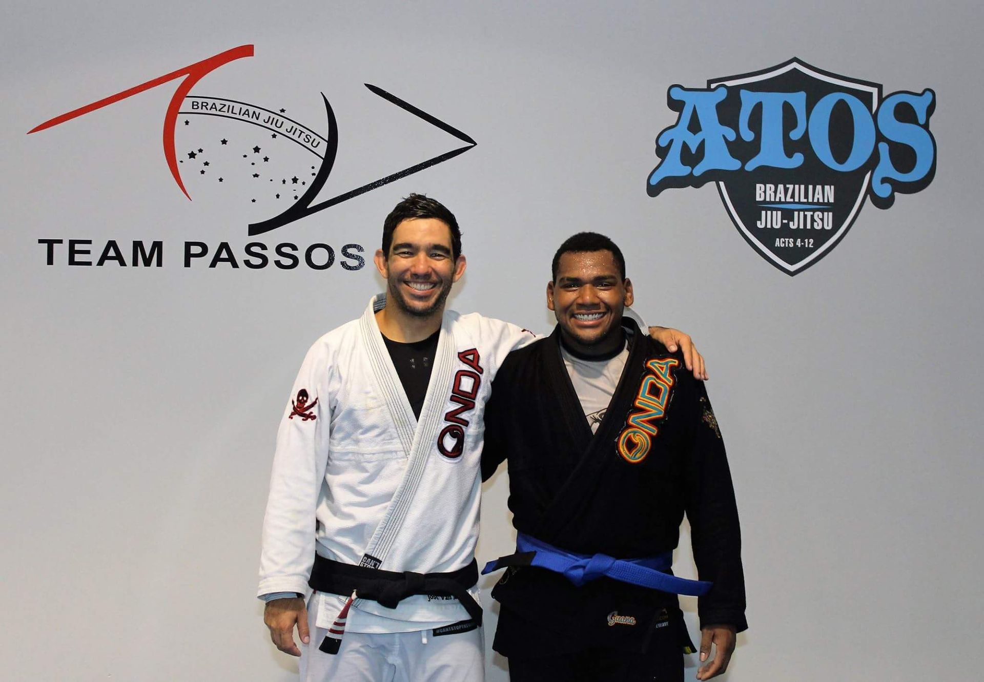 Two men in Jiu-Jitsu uniforms pose with arms around each other in front of logos for Team Passos and ATOS Jiu-Jitsu.