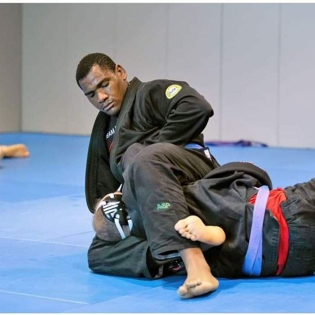 Man in black gi, on top of opponent in a BJJ match, knee on belly. Gym setting, blue mat.