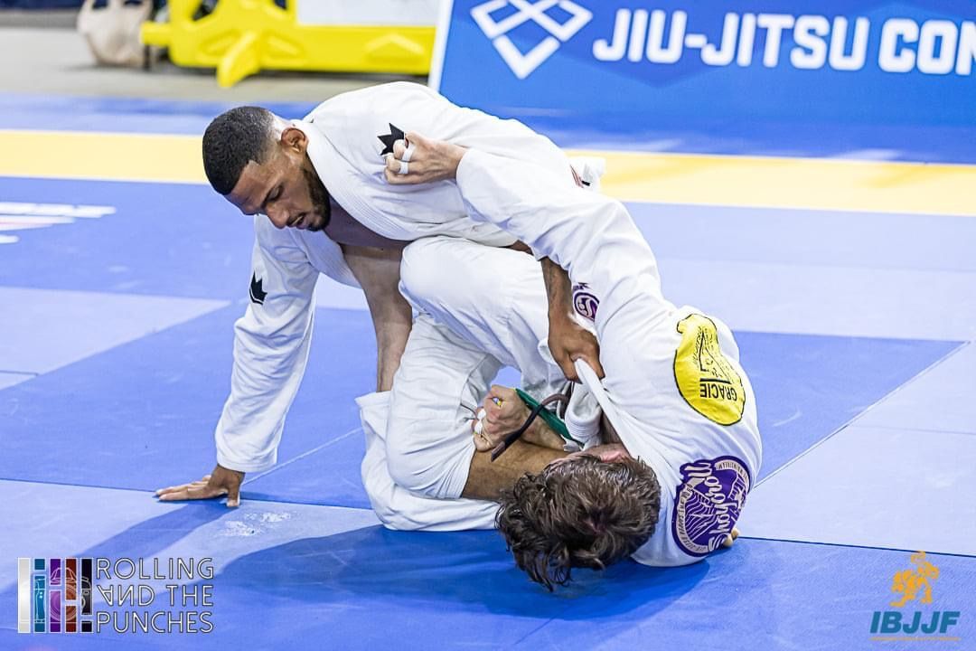 Two men in white gis grapple on a blue mat, one in a top position trying to pass the guard.