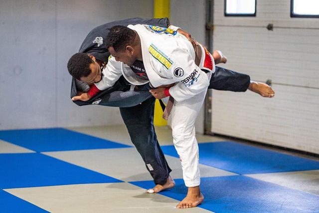 Two men in Jiu-Jitsu uniforms grappling in a gym. One attempts a takedown, lifting the other.