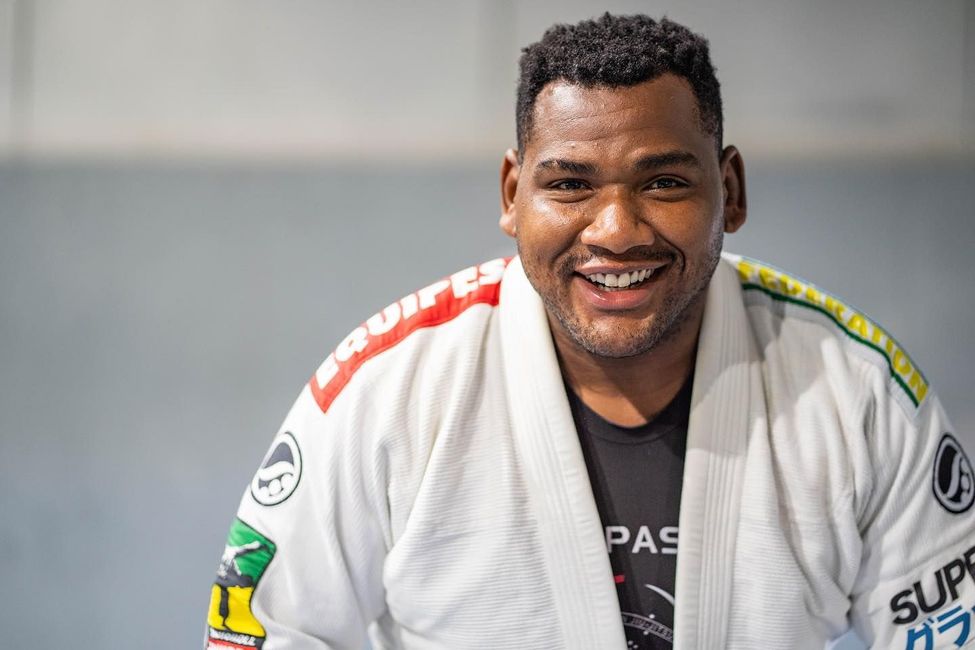 Man in white Jiu-Jitsu uniform smiles; indoor setting with blurred background; logo on gi.