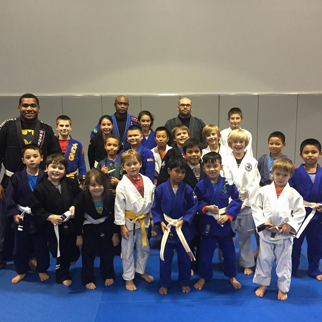 A group of children and instructors in various colored jiu-jitsu uniforms on a blue mat.